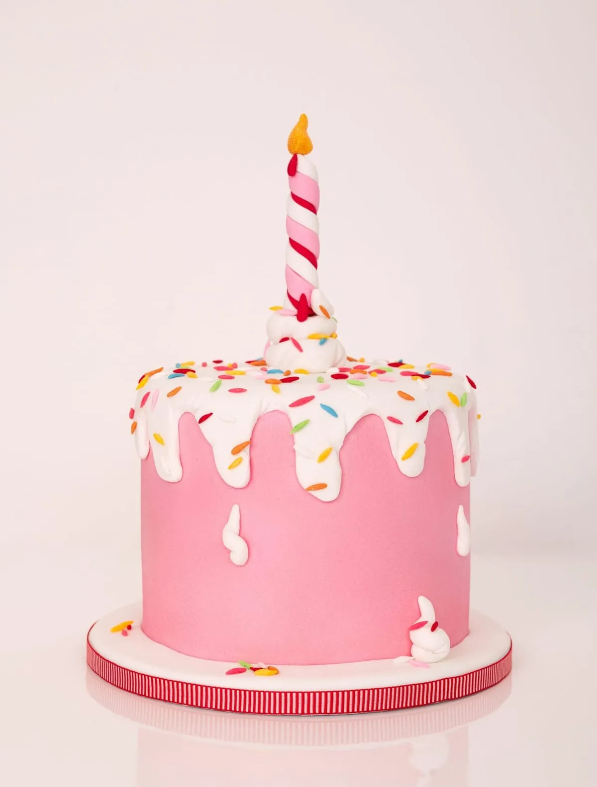 A pink fondant birthday cake with white drips, sprinkles and a candle